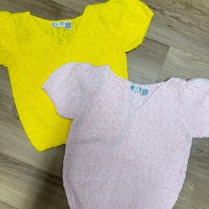 May include: Two knitted sweaters, one yellow and one pink, with short puffed sleeves and a V-neck. Both sweaters have a small tag with the word "Country" on them.