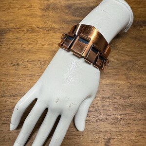 May include: A copper-coloured metal bracelet with a geometric design. The bracelet is composed of rectangular and square shapes. The bracelet is displayed on a white mannequin hand against a wooden background.