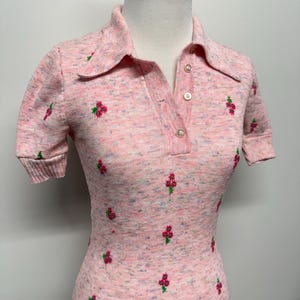 May include: A pink short-sleeved knit shirt with a collared neckline and a button closure. The shirt has a subtle floral pattern with pink and green flowers.