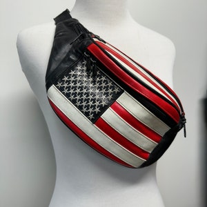May include: A black leather fanny pack with a red, white, and blue American flag design. The flag is embroidered with white stars on a blue field and has red and white stripes.