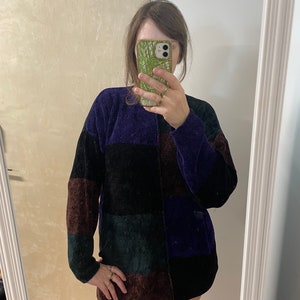 May include: A patchwork sweater in shades of purple, black, green, and brown. The long-sleeved top has a corduroy texture and a relaxed fit. The sweater is reflected in a mirror.