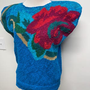 Może przedstawiać: Niebieski sweter z dzianiny z dużym kwiatowym wzorem. Kwiat jest czerwony i magenta z turkusowymi i zielonymi akcentami. Sweter ma złote wykończenia na ramionach i wokół kwiatowego wzoru.