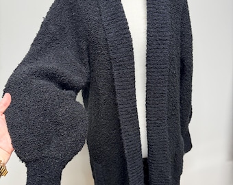 May include: A black, open-front cardigan with a textured, bouclé knit. The long-sleeved sweater features a shawl collar and a relaxed fit. The sleeves have a voluminous, balloon shape, and the cardigan falls to mid-thigh length.