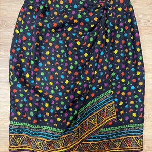 May include: A black wrap skirt with a colorful celestial pattern of stars and crescent moons. The skirt has a decorative border with geometric and floral designs in shades of orange, green, and blue.
