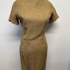 May include: A vintage, knee-length dress in a textured, golden-brown fabric. The dress features short sleeves, a round neckline, and a fitted silhouette with a subtle pocket detail. The dress is displayed on a mannequin.