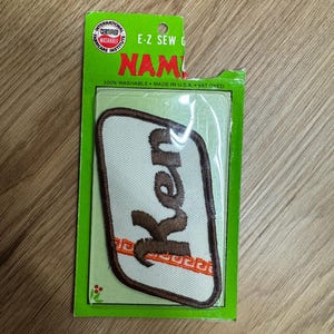 May include: A white iron-on patch with brown lettering that says "Ken" and an orange and white border. The patch is packaged in a clear plastic bag with a green and white cardboard backing that says "E-Z Sew On Name 100% Washable Made in USA. Vat Dyed."