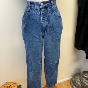 May include: A pair of blue denim jeans with a high waist and a straight leg. The jeans are on a wooden mannequin.