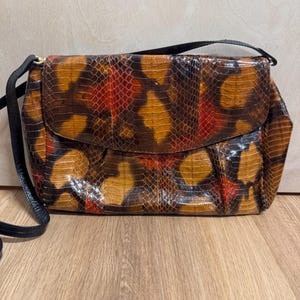 Op de afbeelding: Een klein bruin, rood en zwart schoudertasje van slangenleer met een zwarte band.