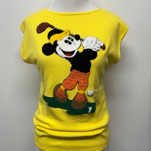 May include: Yellow sleeveless shirt with a large embroidered image of Mickey Mouse playing golf. Mickey is wearing a yellow hat, black shorts, and orange pants. The background is green.
