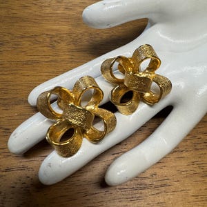 Puede incluir: Un par de pendientes dorados con forma de lazo texturizado. Los pendientes se exhiben sobre una mano blanca sobre un fondo de madera. Los pendientes tienen una superficie texturizada detallada y el diseño del lazo es claramente visible.