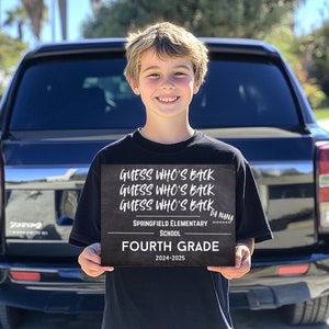 Puede incluir: Un niño sostiene un letrero negro de pizarra que dice "Guess who's back" tres veces, "Springfield Elementary School", "Da Nana", "Fourth Grade" y "2024-2025".
