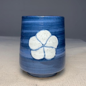 Puede incluir: Una taza de cerámica azul con un diseño de flor blanca. La taza tiene un esmalte texturizado y moteado.