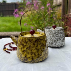 Könnte beinhalten: Zwei Keramik-Teekannen mit goldenen Griffen und Deckeln. Eine ist gelb mit braunen Sprenkeln, die andere weiß mit schwarzen Sprenkeln. Eine Kette aus Bernsteinperlen befindet sich im Vordergrund. Die Teekannen stehen auf einer weißen Oberfläche.