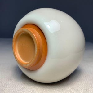 Könnte beinhalten: Ein weißes Keramikobjekt mit abgerundeter Form und einem kontrastierenden orange-braunen Sockel. Der Sockel hat ein kreisförmiges Design mit einer vertieften Innenseite. Das Objekt hat eine glänzende Oberfläche, was auf einen dekorativen oder funktionalen Zweck hindeutet.