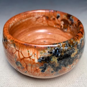 May include: A small, round ceramic bowl with a crackled glaze in shades of orange, brown, and black. The bowl has a slightly uneven rim and a smooth interior.