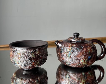 May include: A ceramic tea set featuring a small teapot and a matching cup. The set has a textured, speckled surface with brown, blue, and green hues. The teapot has a lid and a handle. The cup is round with a dark interior.