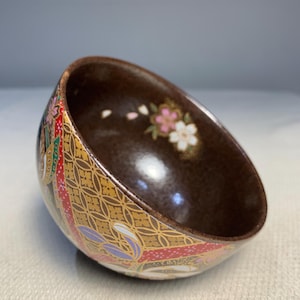 May include: A small, brown ceramic bowl with a gold and white floral design. The bowl has a glossy finish and is decorated with cherry blossoms and other floral motifs.