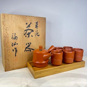 Puede incluir: Un juego de té de color terracota con una tetera, seis tazas y una bandeja de madera. El juego se exhibe frente a una caja de madera con caracteres japoneses. El juego de té es de un tono cálido y terroso.