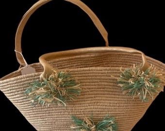 May include: A woven straw tote bag with a curved handle and decorative tassels. The bag features a light brown color with accents of green and tan on the tassels. The handle is a light tan color.