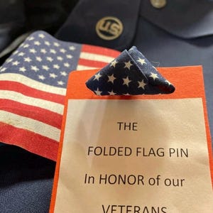 May include: A folded American flag pin with white stars on a blue background and red and white stripes. The pin is on a red card with the text "THE FOLDED FLAG PIN In HONOR of our VETERANS". A US emblem is visible.