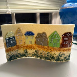 May include: A rectangular glass art piece featuring a row of colorful houses. The houses have various roof designs and colors, including blue, yellow, green, and brown. The base has a textured appearance with sand-like details and green accents.