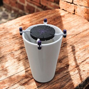 May include: A white ceramic mug with a black and white circular filter attached to the top. The filter is held in place by a white plastic frame with purple accents.