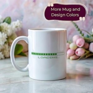 May include: White ceramic mug featuring a green "Loading..." graphic. The mug has a curved handle. The background is blurred with floral elements. A text overlay reads "More Mug and Design Colors".