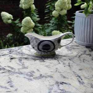 May include: A white ceramic gravy boat with a black and blue circular design and script. The gravy boat has a handle and a yellow base. The background includes a brick wall, white flowers, and a white planter.