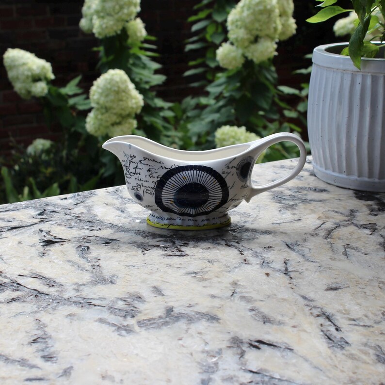 May include: A white ceramic gravy boat with a black and blue circular design and script. The gravy boat has a handle and a yellow base. The background includes a brick wall, white flowers, and a white planter.