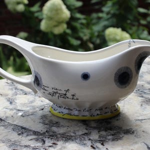 May include: White ceramic gravy boat with a curved handle and spout. The boat features dark blue circular designs and script writing. The base is a bright yellow. A decorative piece for serving sauces and gravies.