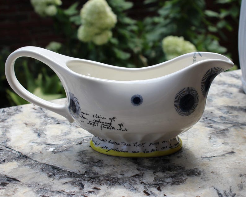 May include: White ceramic gravy boat with a curved handle and spout. The boat features dark blue circular designs and script writing. The base is a bright yellow. A decorative piece for serving sauces and gravies.