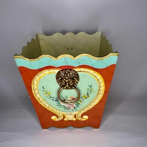 May include: A square planter with a scalloped rim, painted in shades of orange, green, and gold. The planter features a heart-shaped design with a lion's head knocker and floral details.