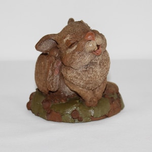 May include: A small, brown ceramic figurine of a rabbit with its eyes closed and mouth open, as if laughing. The rabbit is sitting on a green and brown base with a textured surface.