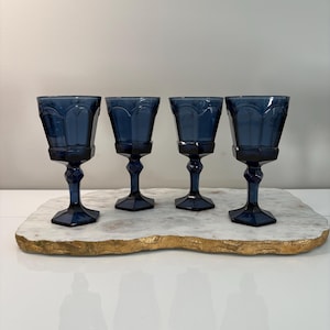 Puede incluir: Cuatro copas de vino de color azul marino con cuencos facetados se colocan sobre una bandeja de mármol blanco con borde dorado. Las copas tienen un diseño clásico y están espaciadas uniformemente.