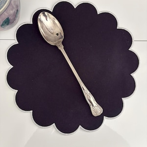 May include: A silver spoon rests on a dark purple, scalloped placemat with a white border. The spoon has an ornate handle design. The placemat is on a white surface. A decorative ceramic pot is partially visible in the background.