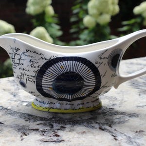 May include: White ceramic gravy boat with a black and blue circular design and handwritten script. The gravy boat has a curved handle and a yellow base. A decorative serving piece.