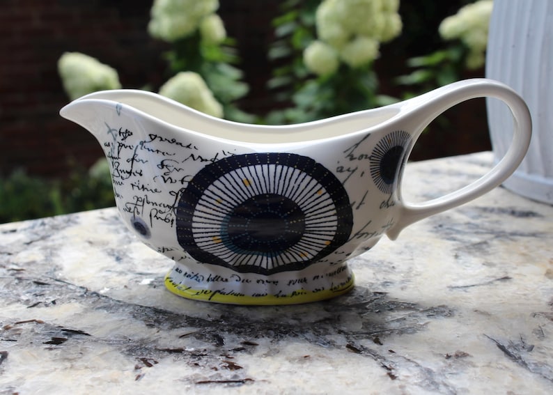 May include: White ceramic gravy boat with a black and blue circular design and handwritten script. The gravy boat has a curved handle and a yellow base. A decorative serving piece.