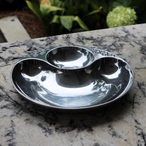 May include: A shiny, oval-shaped, silver serving dish with three connected compartments. The dish has a decorative swirl pattern along the top edge of the two smaller compartments. The surface reflects the surrounding environment.