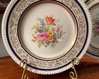 May include: Two white ceramic plates with gold and blue trim and a floral design in the center. The plates are on a gold metal stand.