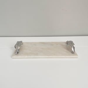 May include: A rectangular marble serving board with a light beige color and subtle veining. It features silver-colored metal handles on each end, adding a decorative touch. The board is designed for serving food.