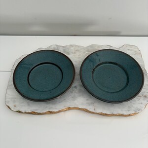 May include: Two teal-colored ceramic saucers with a dark brown rim. The saucers are placed on a white and gold decorative tray. The saucers have a textured surface and a circular indentation in the center.