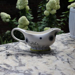 May include: White ceramic gravy boat with a curved handle and spout. The boat features dark blue and gray circular designs and script. The base is accented with a yellow-green band. The gravy boat is on a stone surface.