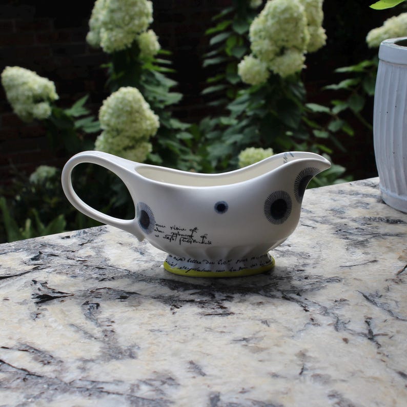 May include: White ceramic gravy boat with a curved handle and spout. The boat features dark blue and gray circular designs and script. The base is accented with a yellow-green band. The gravy boat is on a stone surface.