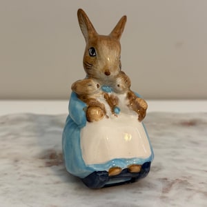May include: A ceramic figurine of a rabbit wearing a blue dress and white apron, holding two smaller rabbits. The figurine has brown and tan fur with black eyes and is a collectible item.