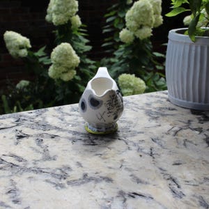 May include: A small, white ceramic creamer with a spout and handle. The creamer features black polka dots and script-like designs. The base has a yellow trim. The creamer is set on a marble-like surface, with greenery in the background.