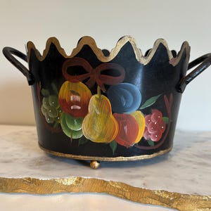 May include: A black metal bowl with a scalloped gold-painted rim and two side handles. The exterior features a hand-painted design of a cluster of colorful fruits, including a yellow pear, red apple, and blue plum, tied together with a brown ribbon.