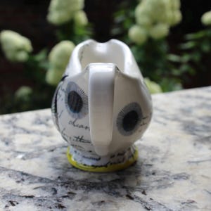 May include: White ceramic creamer with a handle, decorated with stylized eye designs in black and blue. The creamer has a yellow base and script writing. The background is blurred with green foliage.