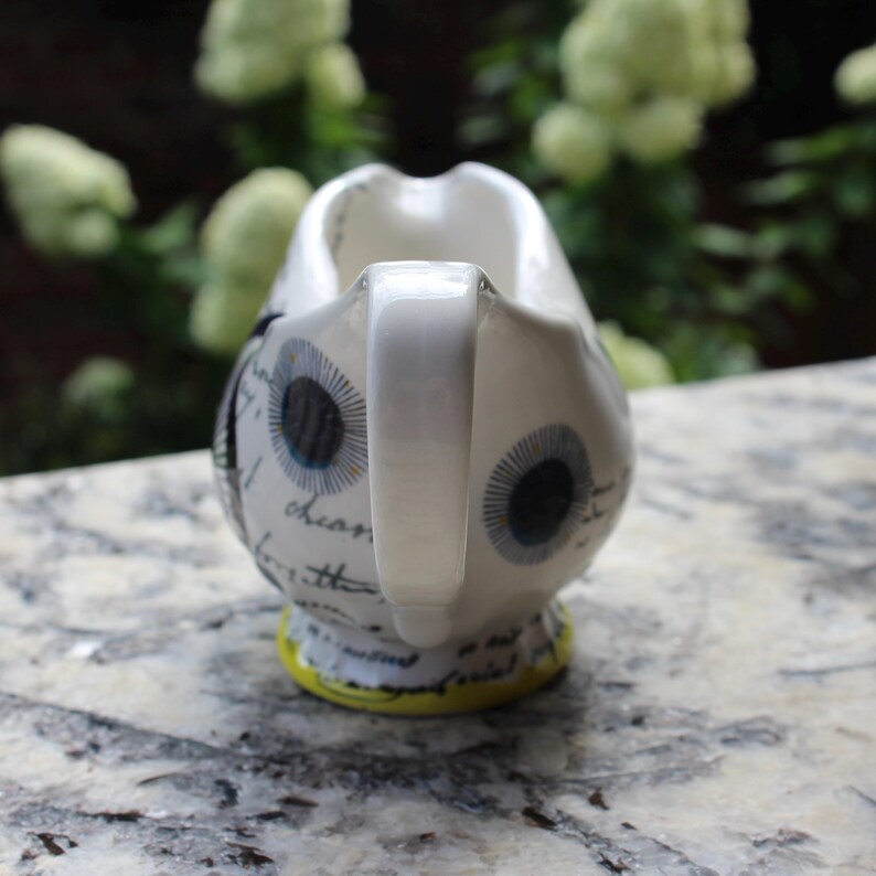May include: White ceramic creamer with a handle, decorated with stylized eye designs in black and blue. The creamer has a yellow base and script writing. The background is blurred with green foliage.