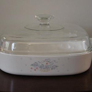 May include: A white casserole dish with a clear glass lid and handle. The dish has a floral design in blue, red, and yellow. The dish is on a dark brown surface.