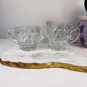 May include: Clear glass sugar bowl and creamer set. The sugar bowl has two handles and a scalloped rim. The creamer has a spout and a handle. Both pieces have a geometric, textured pattern. The set is on a white and gold marble tray.